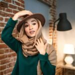 Portrait of a Muslim woman wearing a hijab and hat against a chic indoor brick backdrop.